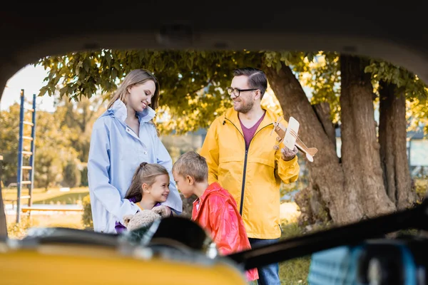 Sonriente hombre sosteniendo juguete cerca de la familia y el coche en primer plano borroso - foto de stock