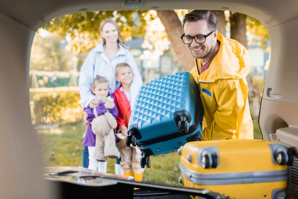 Uomo sorridente in abito autunnale con valigia vicino al bagagliaio di auto e famiglia su sfondo sfocato — Foto stock