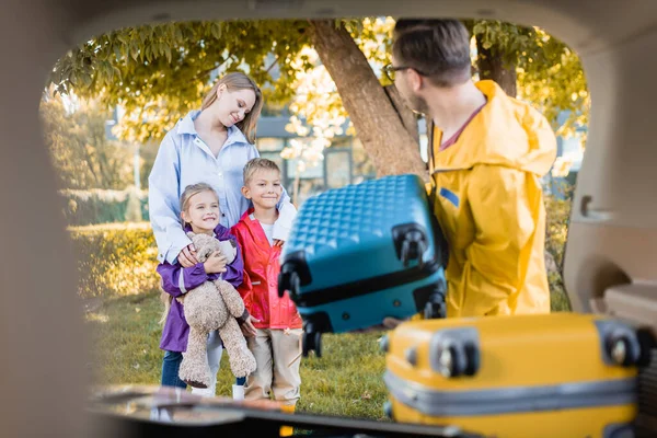 Mulher sorridente abraçando crianças perto do marido segurando mala e porta-malas do carro em primeiro plano borrado — Fotografia de Stock