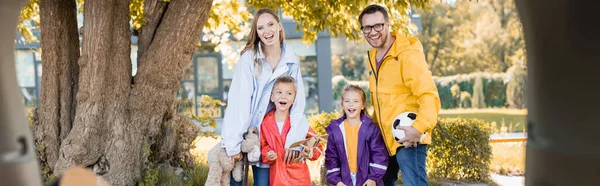 Cheerful family with toys and football standing near car trunk on blurred foreground, banner — Stock Photo