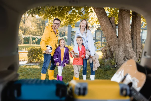Família sorridente em roupa de outono segurando futebol e brinquedos perto do tronco do automóvel em primeiro plano desfocado — Fotografia de Stock