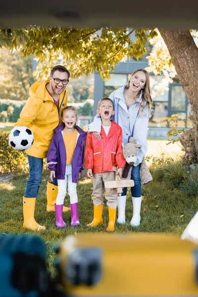 Glückliche Familie im Herbst-Outfit blickt in die Kamera, während sie Fußball und Spielzeug in der Nähe des Autos im verschwommenen Vordergrund hält — Stockfoto
