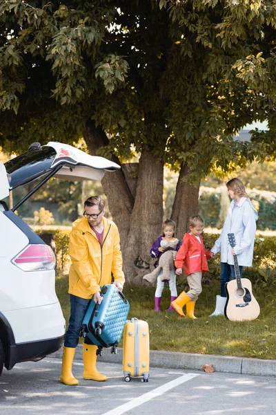 Homem no outono roupa segurando mala perto de carro e esposa com guitarra acústica e crianças no fundo borrado — Fotografia de Stock