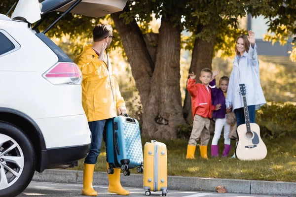 Uomo in abito autunnale agitando mano mentre tiene la valigia vicino auto e moglie con i bambini all'aperto — Foto stock