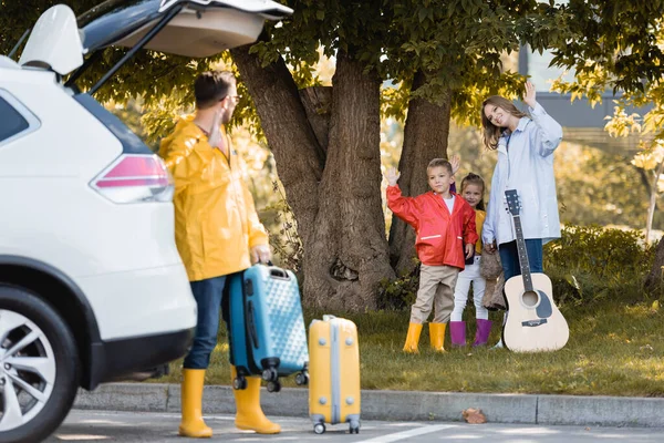 Família em roupa de outono com guitarra acústica acenando para o pai com mala perto do carro em primeiro plano borrado ao ar livre — Fotografia de Stock