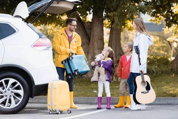 Улыбающийся мужчина в осеннем наряде держит чемодан рядом с семьей с игрушкой и акустической гитарой и авто на открытом воздухе — стоковое фото