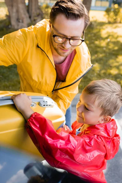Sonriente niño sosteniendo la maleta cerca del padre y el maletero del coche al aire libre - foto de stock