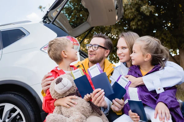Pais sorridentes abraçando crianças com passaportes e passagens aéreas perto do carro ao ar livre — Fotografia de Stock