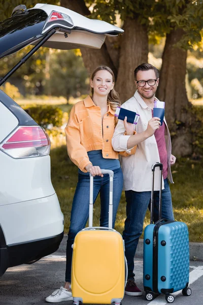 Coppia sorridente con valigie e passaporti guardando la fotocamera vicino all'auto all'aperto — Foto stock