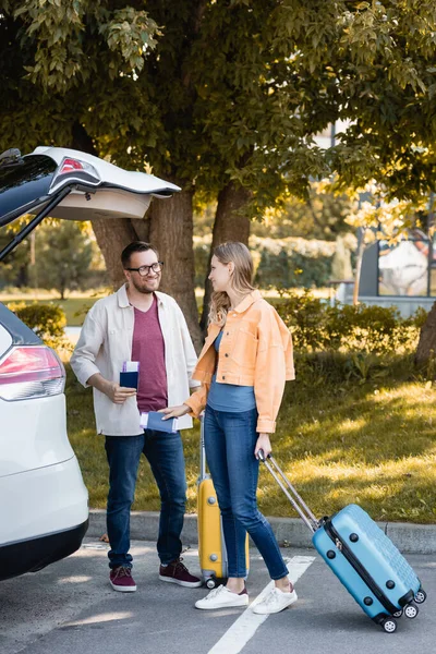 Smiling woman with passport and suitcase looking at husband near auto outdoors — Stock Photo