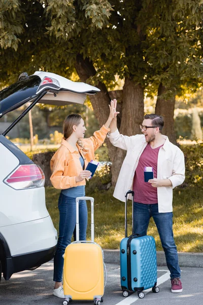 Cheerful couple giving high five while holding passports with air tickets near suitcases and auto — Stock Photo