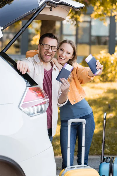 Casal alegre segurando passaportes com passagens aéreas perto de malas e carro ao ar livre — Fotografia de Stock