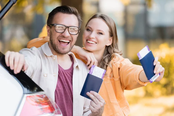 Fröhliche Frau umarmt Ehemann, während sie in der Nähe des Autos Pässe mit Flugtickets in der Hand hält — Stockfoto