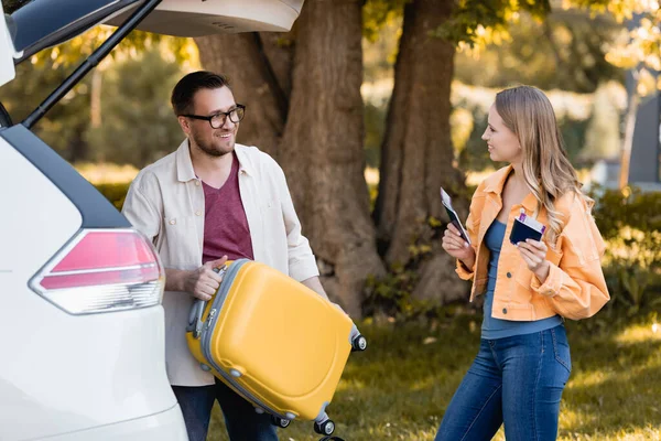 Lächelnde Frau mit Pässen und Flugtickets neben Ehemann mit Koffer und Auto im Freien — Stockfoto