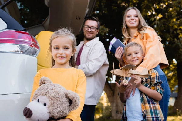 Lächelndes Mädchen mit Spielzeug steht neben Familie mit Pässen und Auto auf verschwommenem Hintergrund — Stockfoto