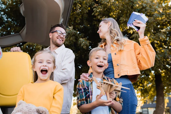 Cheerful kids with toys standing near parents holding passports and suitcase beside car outdoors — Stock Photo