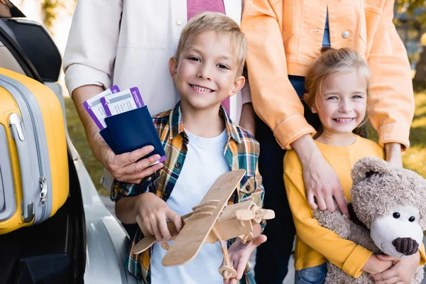 Bambini sorridenti con giocattoli in piedi vicino ai genitori con passaporti e valigia in auto all'aperto — Foto stock