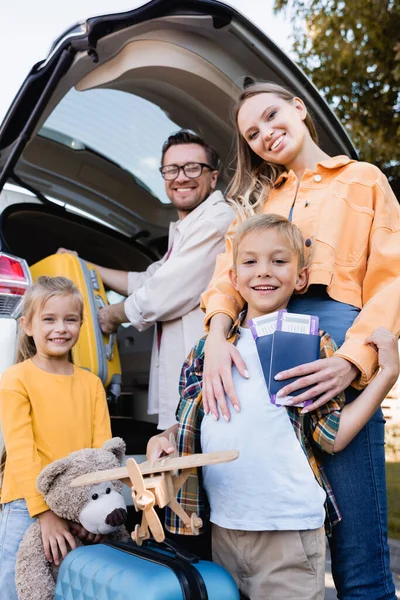 Familia sonriente con pasaportes y maletas mirando la cámara cerca de auto durante el viaje - foto de stock