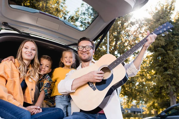 Cheerful family with acoustic guitar looking at camera near truck of auto — Stock Photo