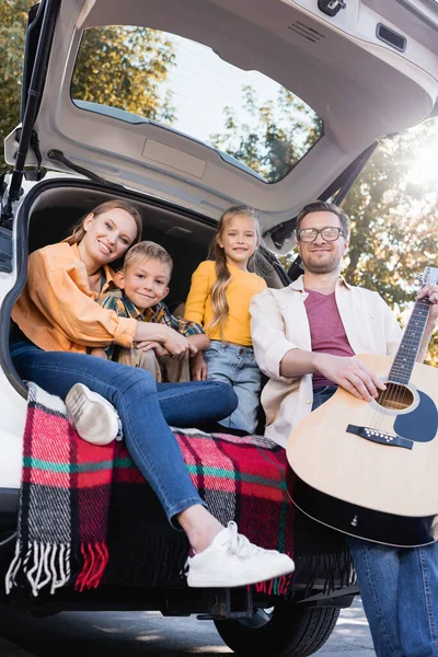 Lächelnde Kinder sitzen neben Eltern mit Akustikgitarre im Kofferraum — Stockfoto