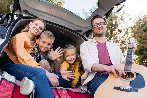 Mann mit Akustikgitarre neben winkenden Kindern und lächelnder Ehefrau im Kofferraum des Autos — Stockfoto