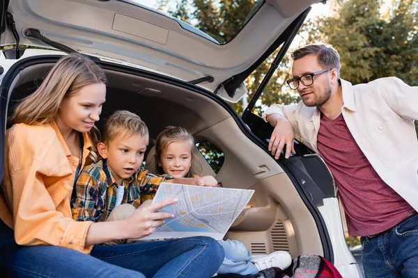 Família sorridente olhando para o mapa perto do tronco do carro durante as férias — Fotografia de Stock