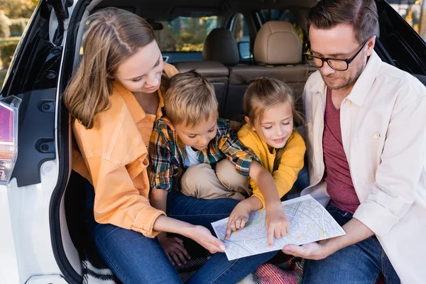 Lächelnde Kinder zeigen mit dem Finger auf Karte in der Nähe von Eltern im Kofferraum — Stockfoto