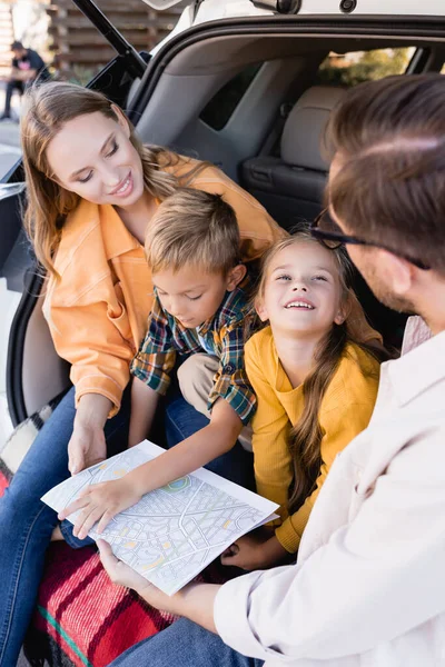 Menina sorridente olhando para o pai perto da família com mapa no porta-malas do carro — Fotografia de Stock