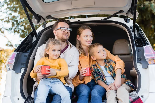 Parents souriants tenant des tasses et embrassant les enfants dans le coffre de la voiture pendant les vacances — Photo de stock