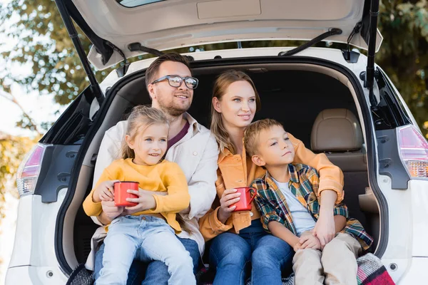 Lächelnde Eltern, die Tassen in der Hand halten und Kinder im Kofferraum umarmen — Stockfoto