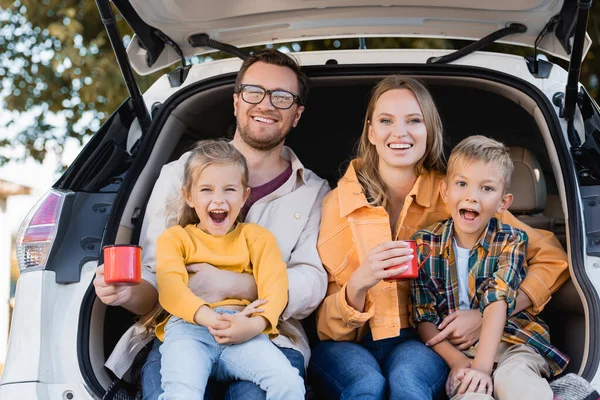 Fröhliche Familie hält Tassen in der Hand und blickt in die Kamera, während sie im Kofferraum des Autos sitzt — Stockfoto