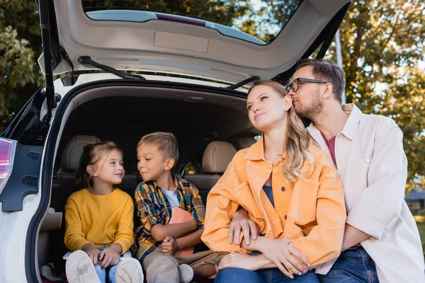 Hombre abrazando esposa cerca sonriendo niños en el maletero del coche - foto de stock