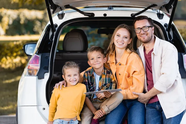 Família alegre com mapa olhando para a câmera perto do tronco do carro ao ar livre — Fotografia de Stock