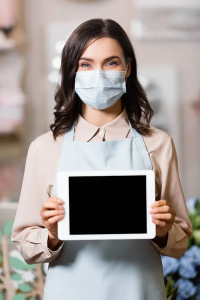 Florista en máscara médica celebración de la tableta con pantalla en blanco mientras mira a la cámara - foto de stock