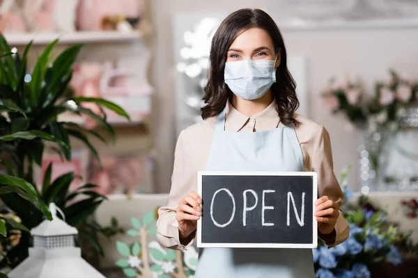 Fiorista positivo in maschera medica tenendo bordo con scritte aperte in negozio di fiori vicino a piante su sfondo sfocato — Foto stock