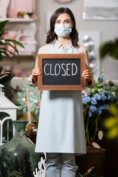 Young florist in medical mask holding board with closed lettering in flower shop on blurred foreground — Stock Photo