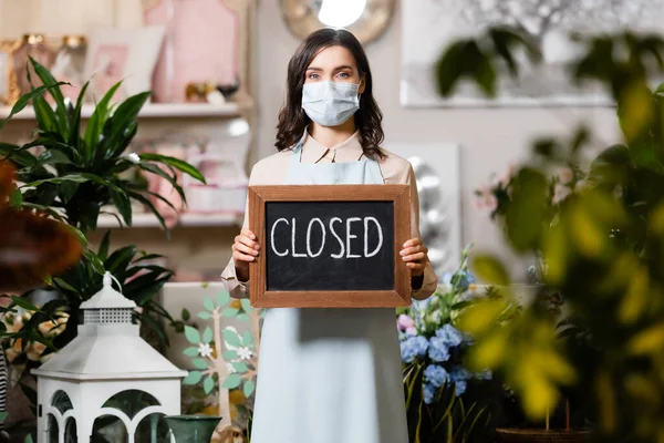 Young florist in medical mask holding board with closed lettering near plants on blurred foreground — Stock Photo