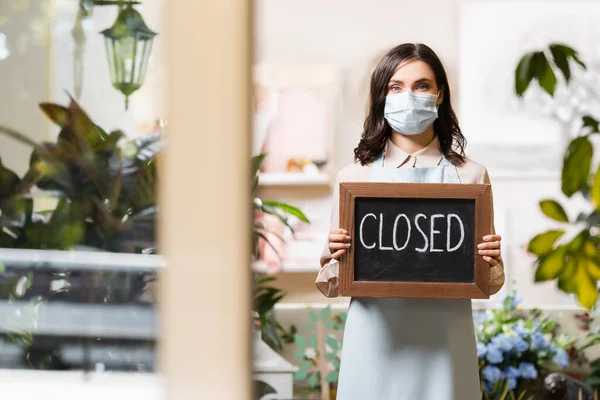 Jovem florista em máscara médica segurando bordo com letras fechadas em primeiro plano borrado — Fotografia de Stock