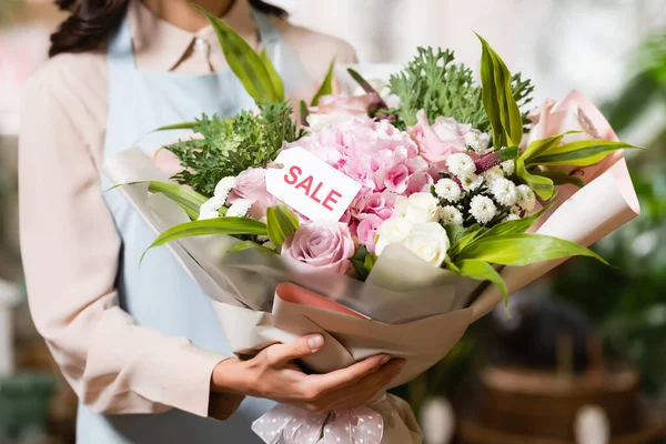 Vista parziale del fiorista che tiene bouquet festivo con lettere di vendita su etichetta su sfondo sfocato — Foto stock