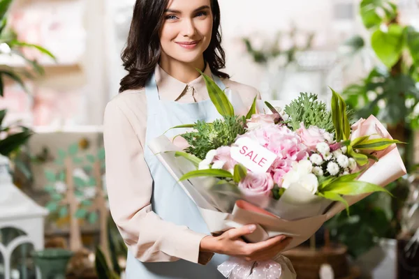 Happy florist holding bouquet with sale lettering on tag while looking at camera near blurred plants on background — Stock Photo