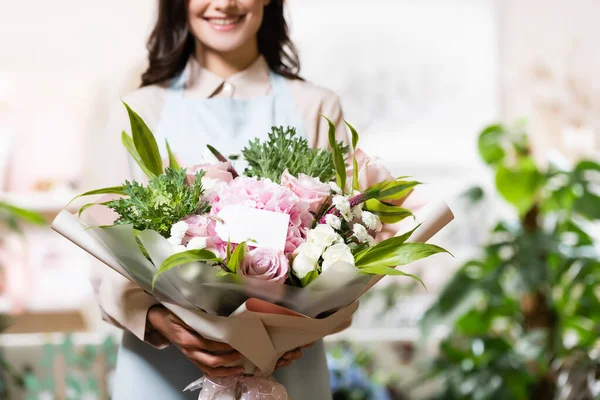 Fiorista sorridente che tiene bouquet con tag vuoto su sfondo sfocato — Foto stock