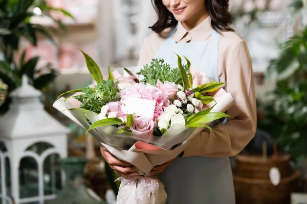 Vista ritagliata di fiorista sorridente che tiene bouquet con tag vuoto vicino alle piante su sfondo sfocato — Foto stock