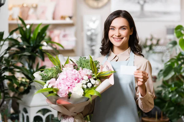 Giovane fiorista con biglietto da visita vuoto e bouquet in negozio di fiori su sfondo sfocato — Foto stock