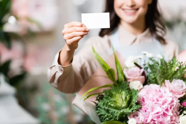 Vista parcial de florista sonriente sosteniendo la tarjeta de visita en blanco y el ramo festivo sobre fondo borroso - foto de stock