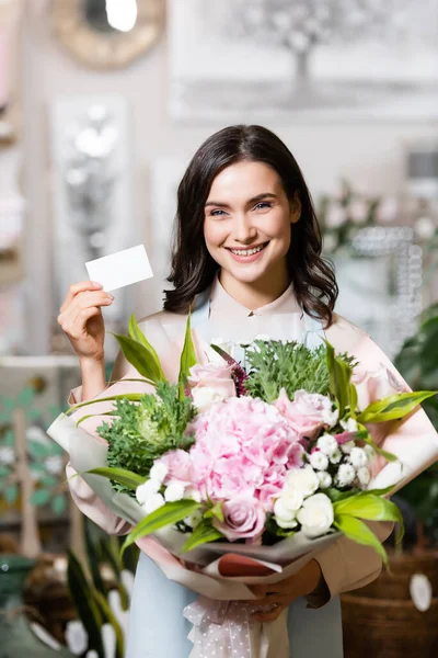 Floristería feliz celebración de la tarjeta de visita en blanco y ramo mientras mira a la cámara en la tienda de flores - foto de stock