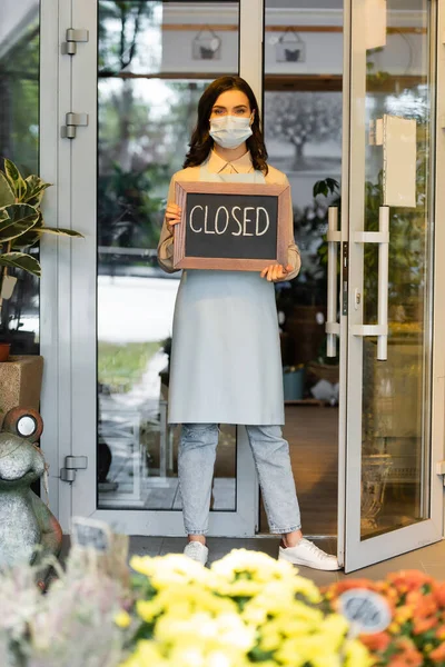 Young florist holding board with closed lettering near entrance of flower shop and flowers on blurred foreground — Stock Photo