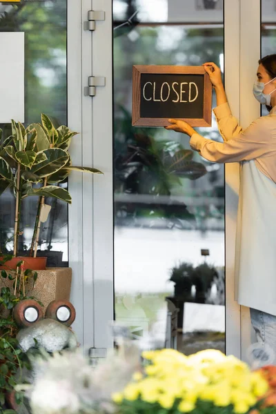 Florista em máscara médica colocando placa com letras fechadas na porta de entrada perto de flores em primeiro plano borrado — Fotografia de Stock