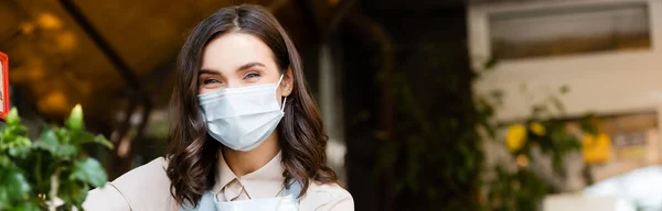 Fiorista positivo in maschera medica guardando la fotocamera nel negozio di fiori su sfondo sfocato, banner — Foto stock