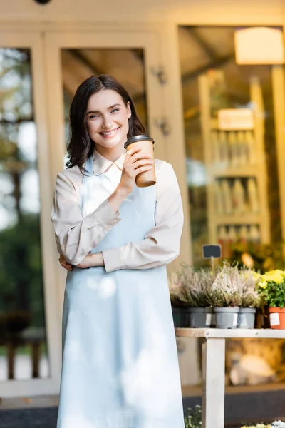 Lächelnder Blumenhändler mit Kaffee in der Nähe von Blumenladen und Topfpflanzen auf verschwommenem Hintergrund — Stockfoto