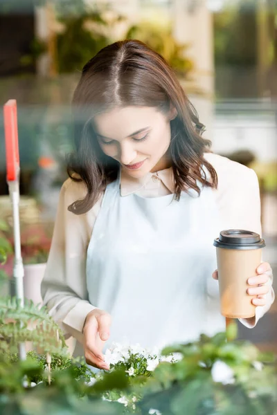 Florista sonriente sosteniendo café para llevar mientras toca flores en primer plano borroso - foto de stock